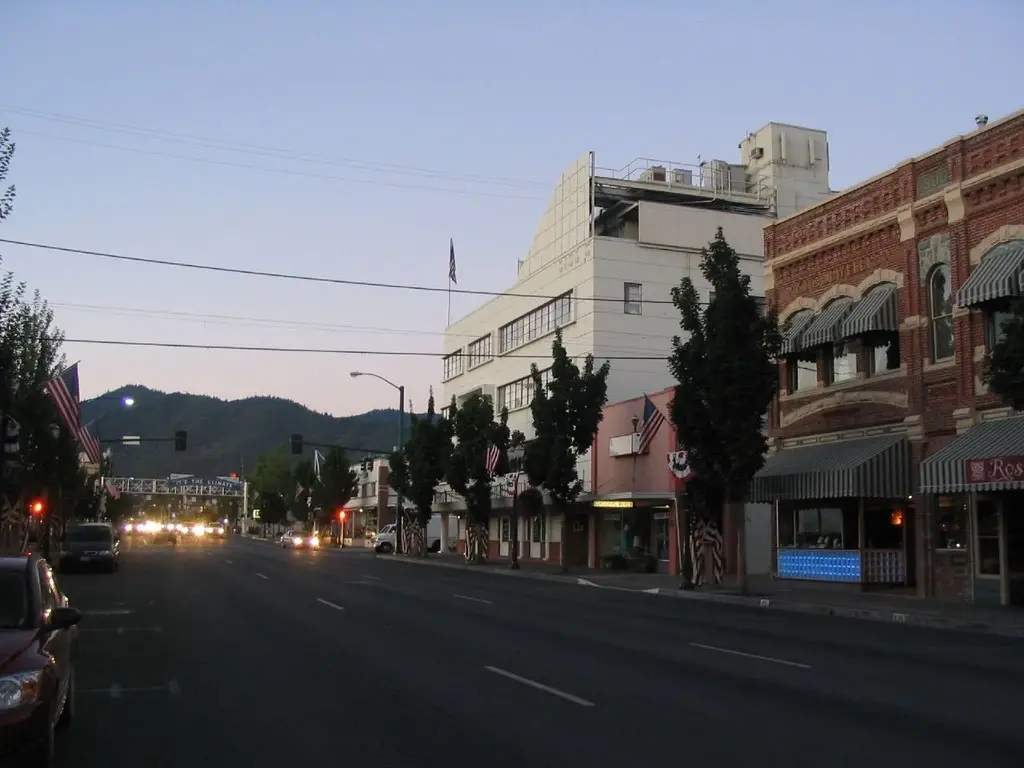 Downtown Grants Pass OR street at dusk with historic brick buildings, modern structure, and mountains in the distance. Local visibility depends on Website Design Grants Pass OR.