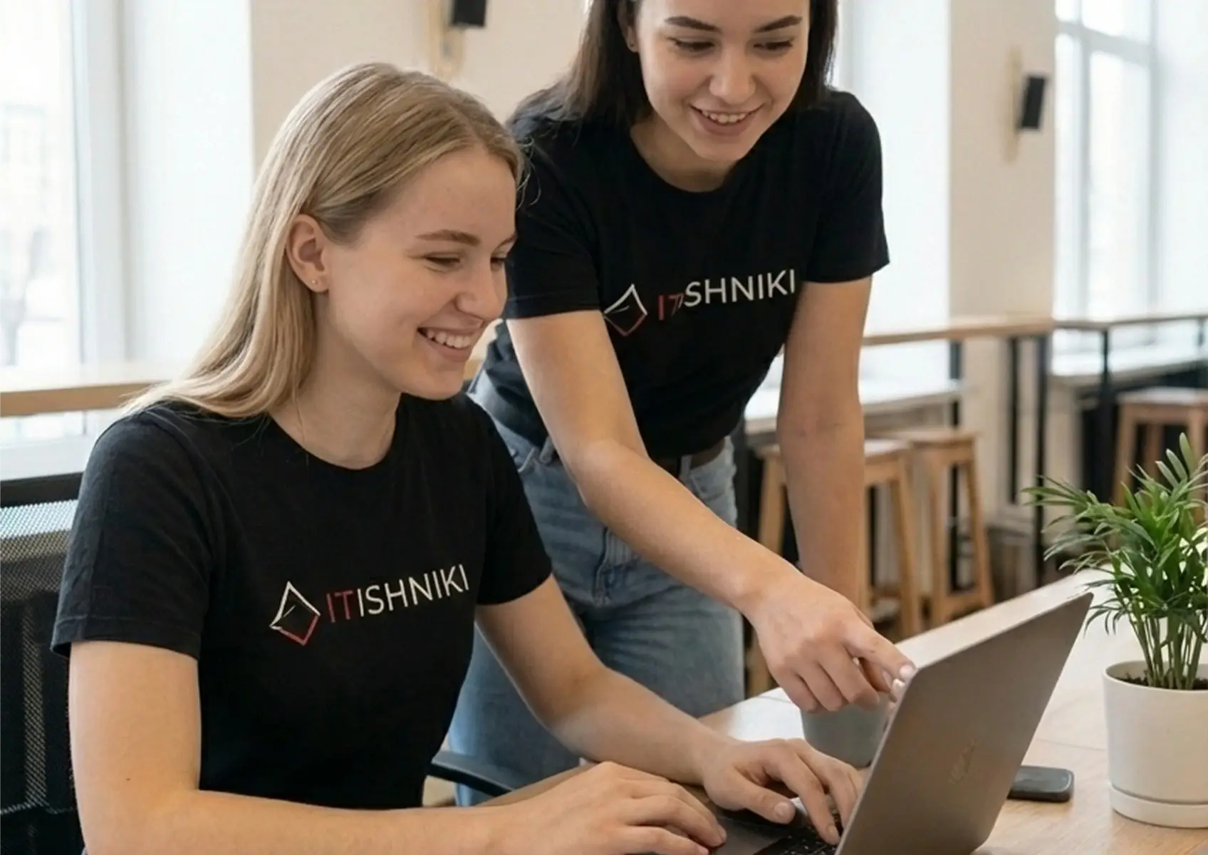 Two smiling women in IT t-shirts reviewing an Elementor template on a laptop in a bright office